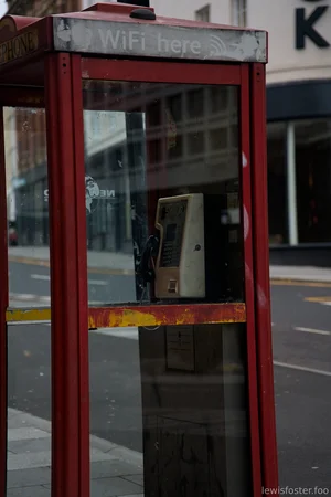 A weathered red public phone box stands empty on a quiet city street, with a faded sign reading “WiFi here” above an old payphone inside.