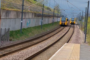 Two Tyne and Wear Metro trains pass each other on parallel tracks, with overhead lines and a concrete wall lining the route.