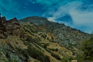 A steep rocky mountainside covered with shrubs and sparse vegetation beneath a vibrant blue sky.