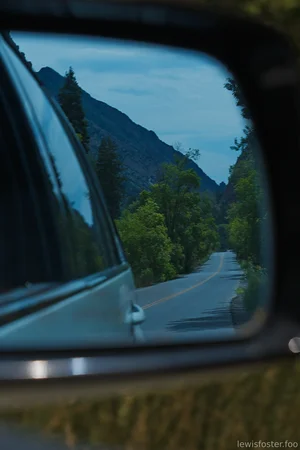 A winding mountain road reflected in a car's side mirror, surrounded by trees and towering peaks.