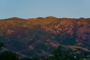 Warm evening sunlight illuminates a rugged mountain ridge with trees and rocky slopes beneath a clear sky.